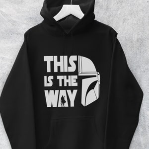 May include: Black hooded sweatshirt with the phrase "THIS IS THE WAY" in white block letters, accompanied by a white helmet graphic and a small silhouette of a figure. The hoodie has a drawstring and a front pocket.