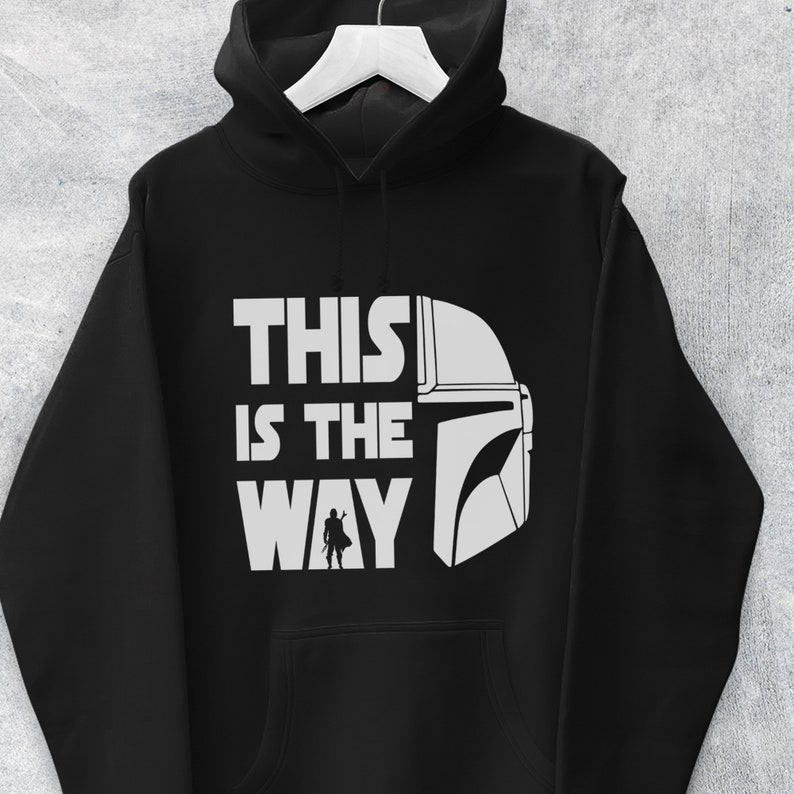 May include: Black hooded sweatshirt with the phrase "THIS IS THE WAY" in white block letters, accompanied by a white helmet graphic and a small silhouette of a figure. The hoodie has a drawstring and a front pocket.