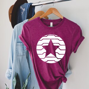 Puede incluir: Una camiseta granate con un dise&ntilde;o de estrella blanca dentro de un c&iacute;rculo blanco. La estrella tiene forma de cinco puntas y el c&iacute;rculo est&aacute; formado por l&iacute;neas horizontales. La camiseta est&aacute; colgada en una percha de madera.