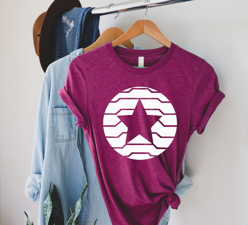 Puede incluir: Una camiseta granate con un dise&ntilde;o de estrella blanca dentro de un c&iacute;rculo blanco. La estrella tiene forma de cinco puntas y el c&iacute;rculo est&aacute; formado por l&iacute;neas horizontales. La camiseta est&aacute; colgada en una percha de madera.