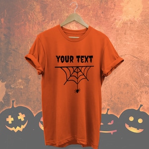 May include: An orange t-shirt with rolled sleeves, displaying the text "YOUR TEXT" above a black spider web design with a hanging spider. The shirt hangs on a wooden hanger against a textured background with pumpkin silhouettes.