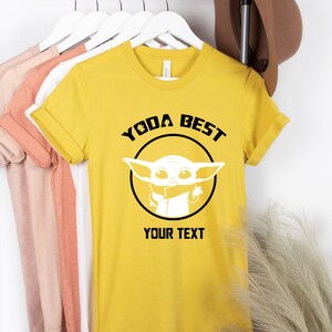 May include: Mustard yellow t-shirt with the text "YODA BEST" above a white graphic of a character from a popular science fiction franchise. The text "YOUR TEXT" is below the graphic. The shirt is hanging on a white hanger.