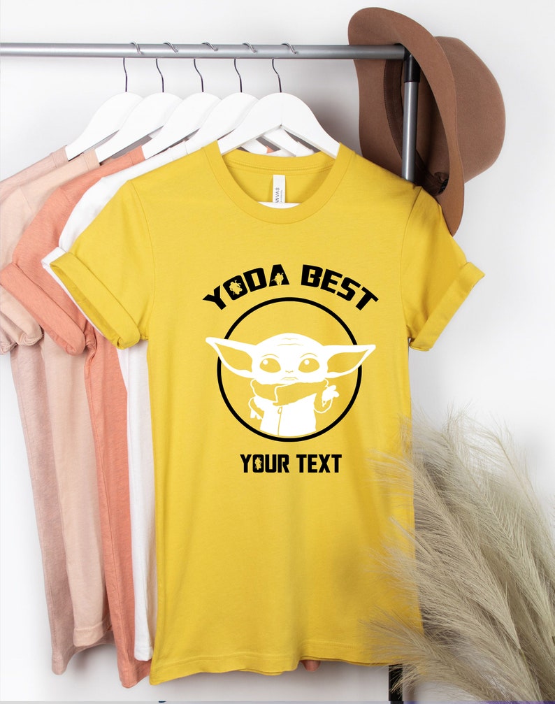 May include: Mustard yellow t-shirt with the text "YODA BEST" above a white graphic of a character from a popular science fiction franchise. The text "YOUR TEXT" is below the graphic. The shirt is hanging on a white hanger.
