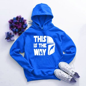 May include: A royal blue hooded sweatshirt with the text "THIS IS THE WAY" in white, along with a white helmet graphic. The image also includes white sneakers, a purple floral arrangement, and a small gift box.