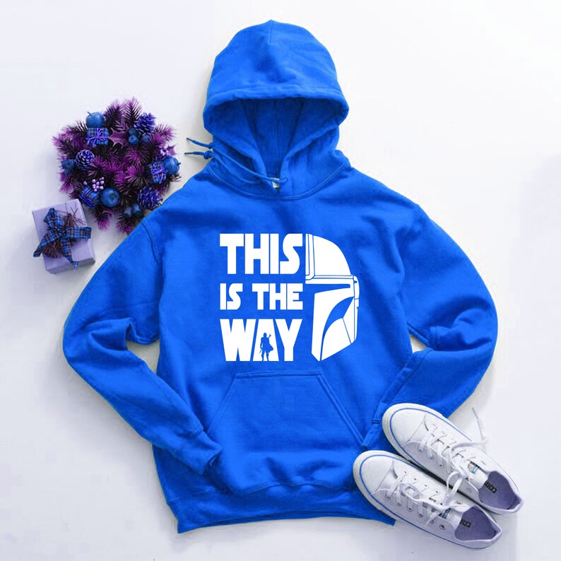 May include: A royal blue hooded sweatshirt with the text "THIS IS THE WAY" in white, along with a white helmet graphic. The image also includes white sneakers, a purple floral arrangement, and a small gift box.