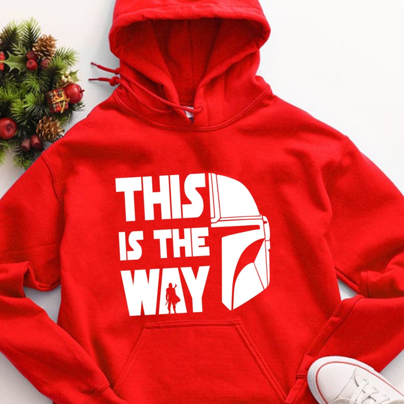 May include: A red hooded sweatshirt with the phrase "THIS IS THE WAY" in white block letters, accompanied by a white graphic of a helmet and a small figure. The sweatshirt has a front pocket and a drawstring hood.