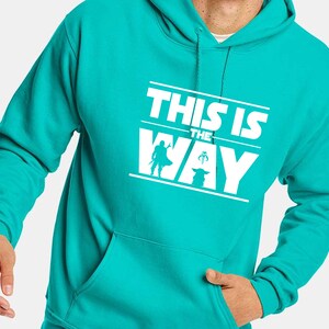 Puede incluir: Sudadera con capucha turquesa con la frase blanca "THIS IS THE WAY" y siluetas de personajes. La sudadera tiene un bolsillo delantero y una capucha con cord&oacute;n.