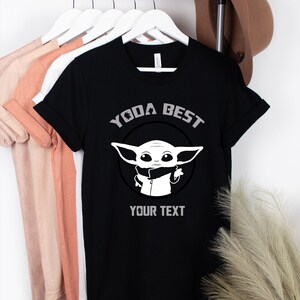 May include: Black t-shirt with the text "YODA BEST" and a graphic of a cartoon character. The shirt also has the text "YOUR TEXT" below the graphic. The shirt is hanging on a white hanger with other shirts.