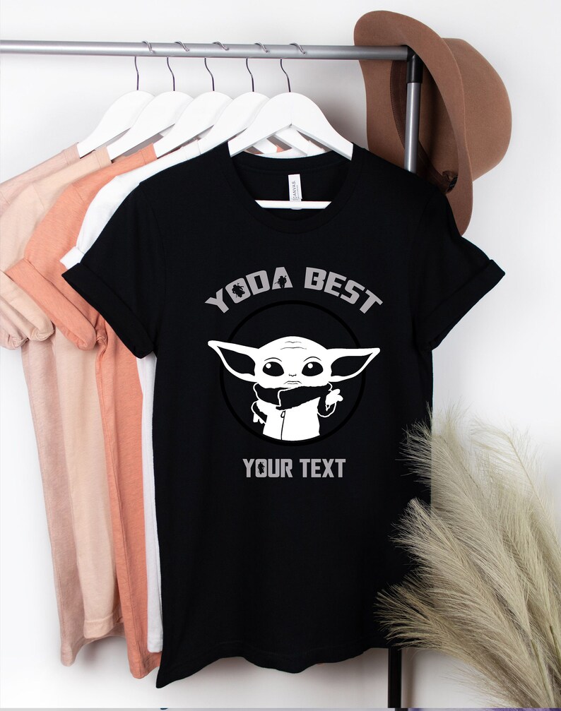 May include: Black t-shirt with the text "YODA BEST" and a graphic of a cartoon character. The shirt also has the text "YOUR TEXT" below the graphic. The shirt is hanging on a white hanger with other shirts.