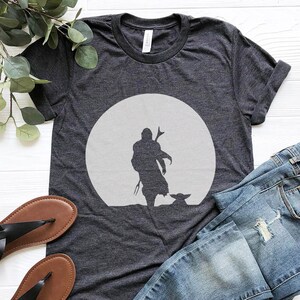 May include: Dark gray t-shirt featuring a silhouette of a figure with a child-like character in front of a large, light gray circle. The shirt is paired with blue jeans and black sandals, suggesting casual wear.
