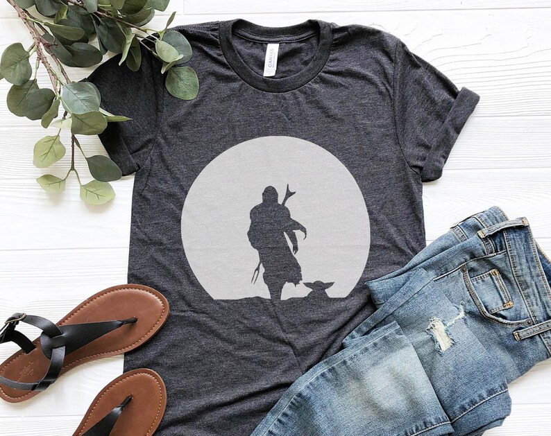 May include: Dark gray t-shirt featuring a silhouette of a figure with a child-like character in front of a large, light gray circle. The shirt is paired with blue jeans and black sandals, suggesting casual wear.