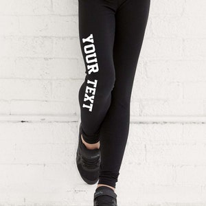 May include: Black leggings with the text "YOUR TEXT" printed vertically in white block letters. The person is wearing black trainers. The background is a white brick wall. The leggings are likely made of a stretchy material.