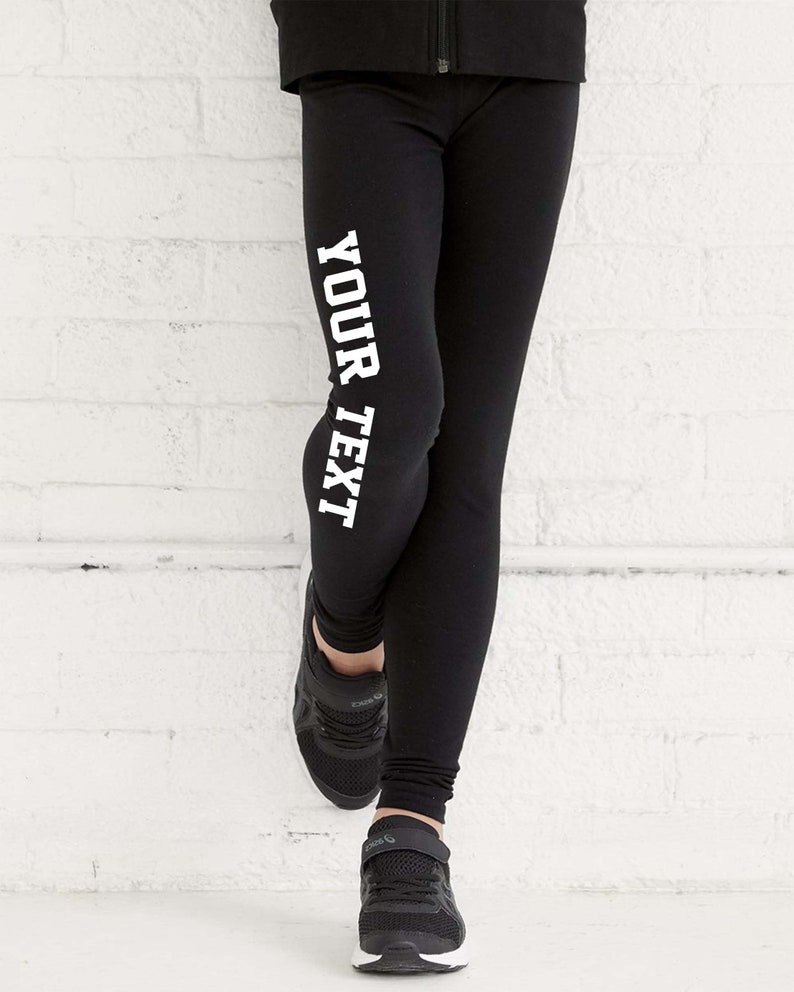 May include: Black leggings with the text "YOUR TEXT" printed vertically in white block letters. The person is wearing black trainers. The background is a white brick wall. The leggings are likely made of a stretchy material.