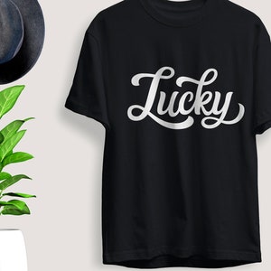 May include: Black t-shirt with the word "Lucky" in white, cursive lettering. The shirt is displayed on a light background, with a dark blue hat and a potted plant with green leaves in the background.