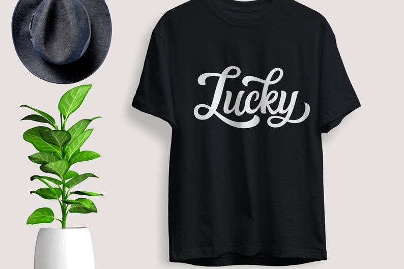 May include: Black t-shirt with the word "Lucky" in white, cursive lettering. The shirt is displayed on a light background, with a dark blue hat and a potted plant with green leaves in the background.