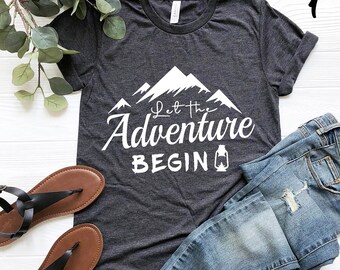 Könnte beinhalten: Dunkelgraues T-Shirt mit dem Text "Let the Adventure Begin" und einem Bergmotiv. Das Shirt wird mit blauen Jeans und braunen Sandalen kombiniert. Das Bild ist auf einer weißen Holzoberfläche drapiert.