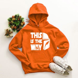 May include: Orange hooded sweatshirt with the text "THIS IS THE WAY" and a white graphic of a helmet and character. White sneakers are placed below the sweatshirt. A small decorative wreath and gift box are to the left.