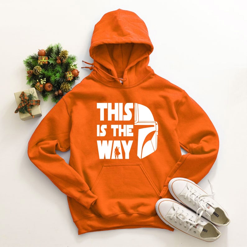May include: Orange hooded sweatshirt with the text "THIS IS THE WAY" and a white graphic of a helmet and character. White sneakers are placed below the sweatshirt. A small decorative wreath and gift box are to the left.