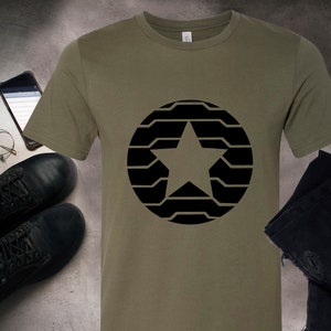 Puede incluir: Camiseta verde oliva con un dise&ntilde;o de estrella negra dentro de un c&iacute;rculo. La camiseta se muestra con botas negras, un reloj, gafas, un tel&eacute;fono inteligente y vaqueros negros. El dise&ntilde;o est&aacute; centrado en la camiseta.