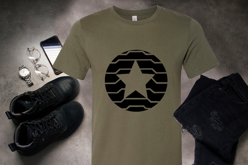 Puede incluir: Camiseta verde oliva con un dise&ntilde;o de estrella negra dentro de un c&iacute;rculo. La camiseta se muestra con botas negras, un reloj, gafas, un tel&eacute;fono inteligente y vaqueros negros. El dise&ntilde;o est&aacute; centrado en la camiseta.