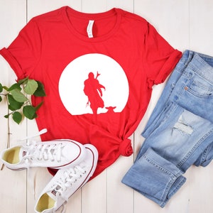 May include: A red t-shirt featuring a white silhouette of a figure and a small creature against a white circle. The shirt is paired with light blue denim jeans and white sneakers.
