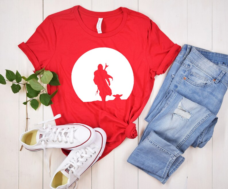 May include: A red t-shirt featuring a white silhouette of a figure and a small creature against a white circle. The shirt is paired with light blue denim jeans and white sneakers.