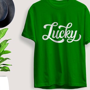 May include: A bright green t-shirt with the word "Lucky" in white script. The shirt is displayed with a dark gray hat and a potted plant with green leaves.