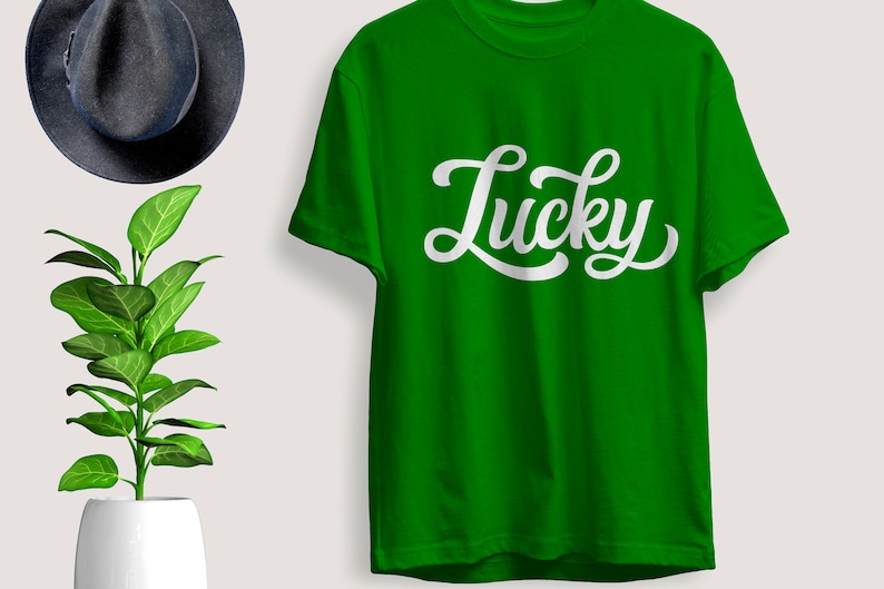 May include: A bright green t-shirt with the word "Lucky" in white script. The shirt is displayed with a dark gray hat and a potted plant with green leaves.