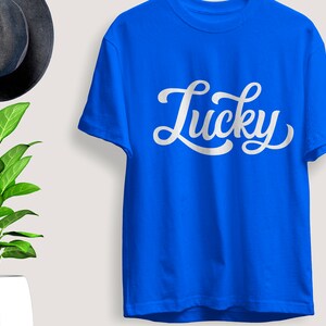 May include: A royal blue t-shirt with the word "Lucky" in white script. The shirt is hanging on a wall next to a dark gray hat and a potted plant with green leaves.