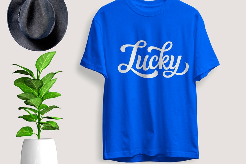 May include: A royal blue t-shirt with the word "Lucky" in white script. The shirt is hanging on a wall next to a dark gray hat and a potted plant with green leaves.