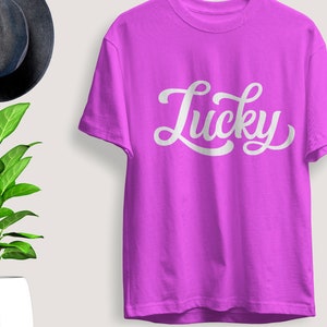 May include: A vibrant purple t-shirt with the word "Lucky" in white script. The shirt is displayed on a light background, with a dark gray hat and a potted plant with green leaves in the background. The t-shirt is a casual wear item.
