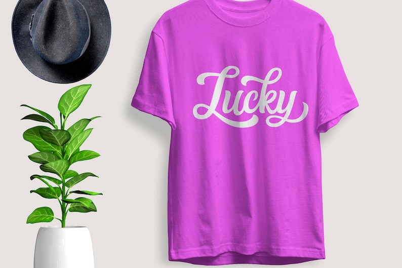 May include: A vibrant purple t-shirt with the word "Lucky" in white script. The shirt is displayed on a light background, with a dark gray hat and a potted plant with green leaves in the background. The t-shirt is a casual wear item.