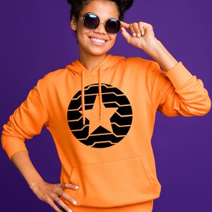 K&ouml;nnte beinhalten: Ein orangefarbener Kapuzenpullover mit einem schwarzen und orangefarbenen Sterndesign. Der Kapuzenpullover hat eine Vordertasche und eine Kapuze. Die Person auf dem Bild tr&auml;gt eine runde Sonnenbrille und l&auml;chelt. Der Hintergrund ist einfarbig lila.