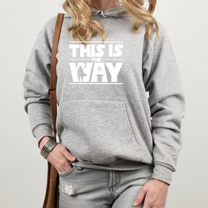 Puede incluir: Una sudadera con capucha gris con un bolsillo delantero y texto blanco que dice "THIS IS THE WAY" con un gr&aacute;fico de un personaje y un beb&eacute;. La sudadera es de estilo casual.