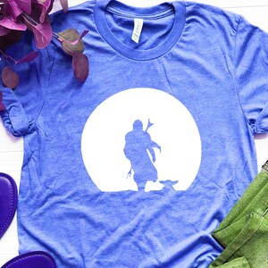 May include: Blue t-shirt featuring a white silhouette of a figure with a weapon, and a small creature, set against a white circle. The shirt is made of a soft material and has a crew neck.