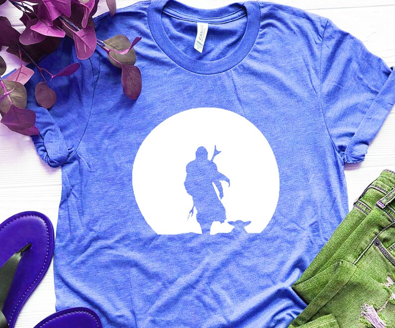 May include: Blue t-shirt featuring a white silhouette of a figure with a weapon, and a small creature, set against a white circle. The shirt is made of a soft material and has a crew neck.