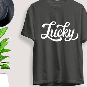 May include: Dark gray t-shirt with the word "Lucky" in white, cursive lettering. The shirt is displayed next to a dark blue hat and a potted plant with green leaves.
