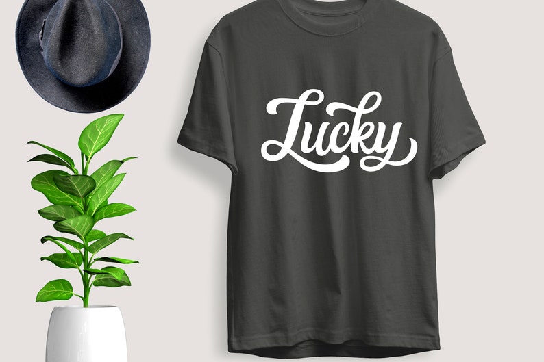 May include: Dark gray t-shirt with the word "Lucky" in white, cursive lettering. The shirt is displayed next to a dark blue hat and a potted plant with green leaves.