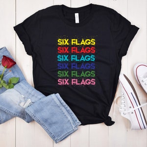 Könnte beinhalten: Schwarzes T-Shirt mit dem Schriftzug "SIX FLAGS" in einem bunten Graffiti-Design. Dazu blaue Jeans, eine rote Rose und weiße Turnschuhe.