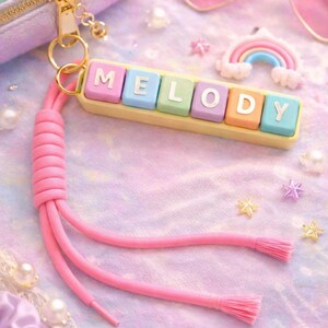 Peut inclure: Image sur le thème pastel avec un porte-clés épelant "MELODY" en blocs de lettres colorés. Le porte-clés a un cordon rose et se trouve à côté d'une pochette arc-en-ciel, de lunettes de soleil en forme de cœur et d'étoiles et de perles décoratives. Le fond est un tissu doux multicolore.