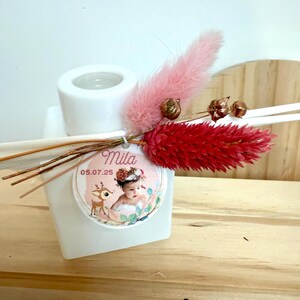 Op de afbeelding: Witte vierkante diffuserfles met een ronde opening, versierd met roze en rode gedroogde bloemen en decoratieve stokjes. Een rond label met de naam "Mila" en de datum "05.07.25" is bevestigd. Het label toont een baby en een hertenillustratie.