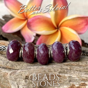 Pode incluir: Close-up de um fio de contas facetadas, vermelho escuro, com detalhes prateados. As contas são exibidas em uma superfície de madeira, com um fundo desfocado de uma flor rosa e amarela. O texto "BEADS & STONES" é visível.