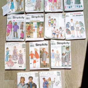 May include: A collection of vintage sewing patterns for children's clothing, featuring various designs and brands like Simplicity and McCall's. The patterns showcase illustrations of dresses, tops, and pants. The patterns are in their original packaging.