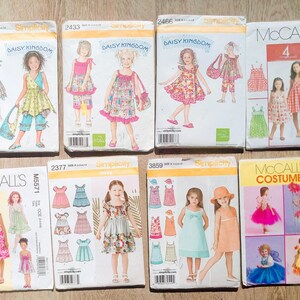 May include: A collection of vintage sewing patterns for children's clothing. The patterns feature illustrations of girls in dresses, jumpsuits, and costumes. Brands include Simplicity and McCall's. The patterns are in various sizes and styles.