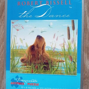 May include: A rectangular puzzle box with a blue border features the text "ROBERT BISSELL the Dance." The puzzle image shows a bear in a marshy landscape with dragonflies. The puzzle is 10 x 8 inches and has 100 interlocking pieces.