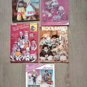 May include: A collection of five vintage sewing pattern books featuring various doll and toy designs. The books have colorful covers with illustrations of the projects, including a patchwork doll, a sock monkey, and a variety of doll clothes.
