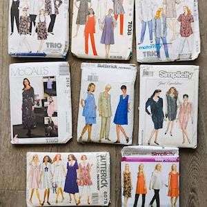 May include: A collection of vintage sewing patterns from McCall's, Butterick, and Simplicity. The patterns feature illustrations of various clothing styles, including dresses, tops, and pants. The patterns are displayed on a wooden surface.