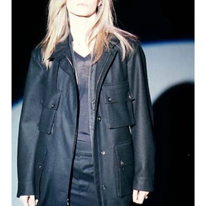 May include: A woman wearing a black long-sleeved jacket with large pockets and a black skirt. The jacket has a zipper and buttons. The woman has long blonde hair.