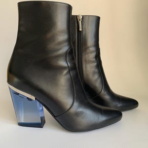 May include: A pair of black leather ankle boots with a pointed toe and a silver zipper. The boots feature a unique heel design with a clear blue acrylic block and silver accents.