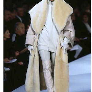 Puede incluir: Una modelo en una pasarela luce un abrigo largo color crema con un gran cuello esponjoso. El abrigo se combina con un atuendo blanco, guantes y botas altas hasta la rodilla. El look general es un conjunto de alta costura con temática invernal.